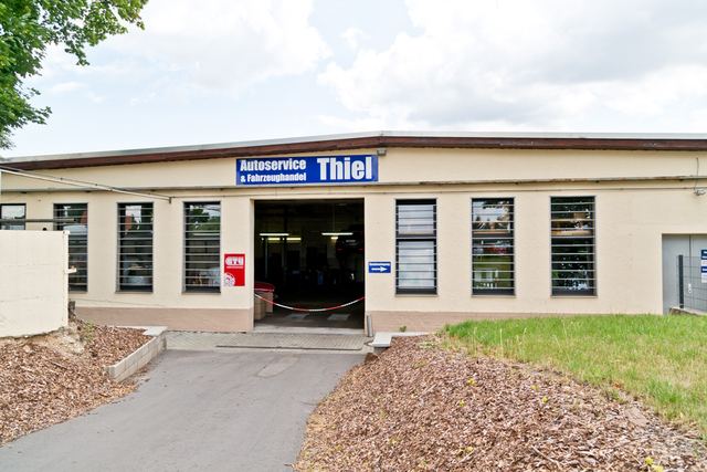 Werkstattgebäude mit dem Schild „Autoservice & Fahrzeughandel Thiel“, offenem Tor und Hinweisschild zum Kundeneingang